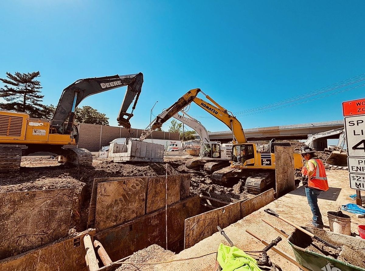 Excavators setting a precast concrete section beside a trench braced with trench boxes along the I-294 Tollway, with a JLA & Sons worker standing at the edge.
