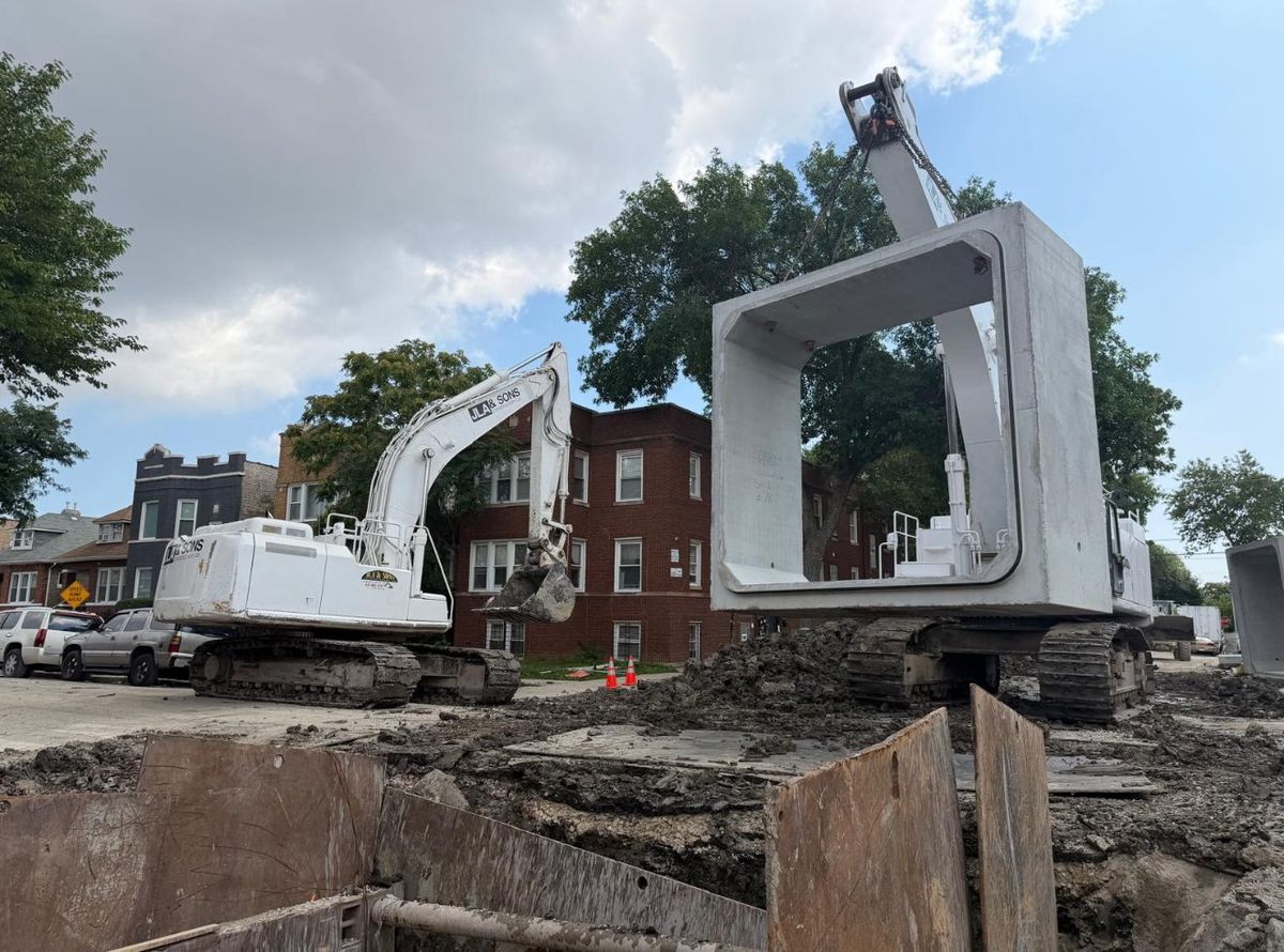 A JLA & Sons excavator lifting a 12-foot square precast concrete box culvert section over a shored trench on W. Le Moyne St, with a second JLA excavator behind it.