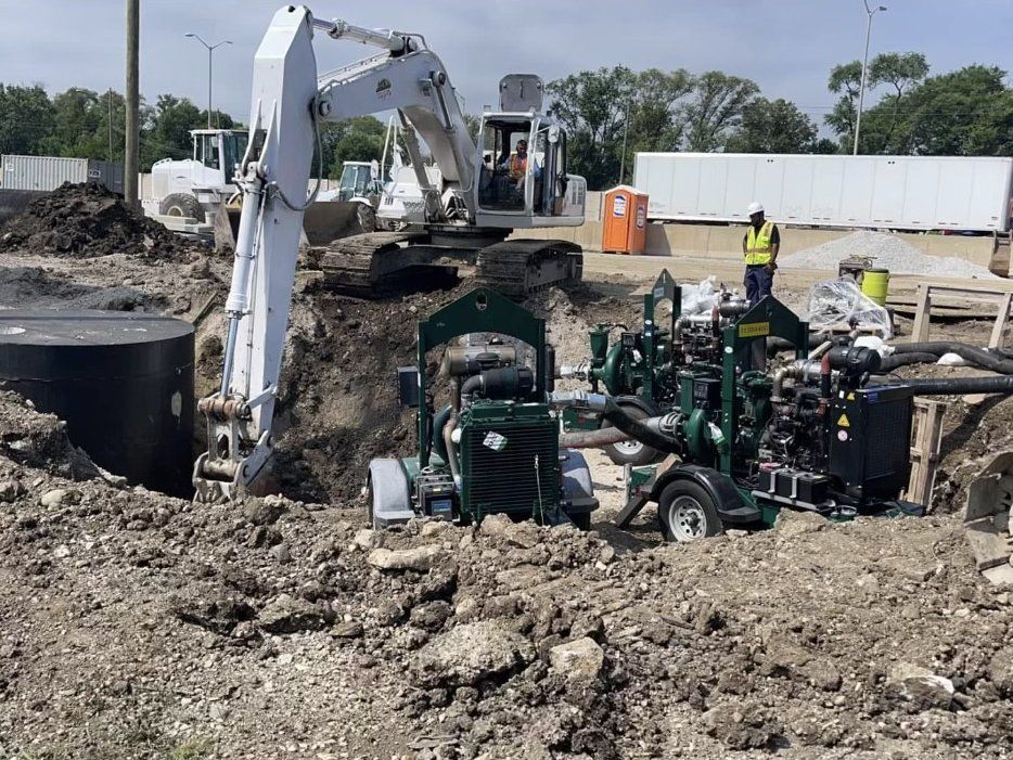 Green diesel bypass pumps and hoses beside an open excavation on an Illinois Tollway job, with a JLA & Sons excavator behind them.