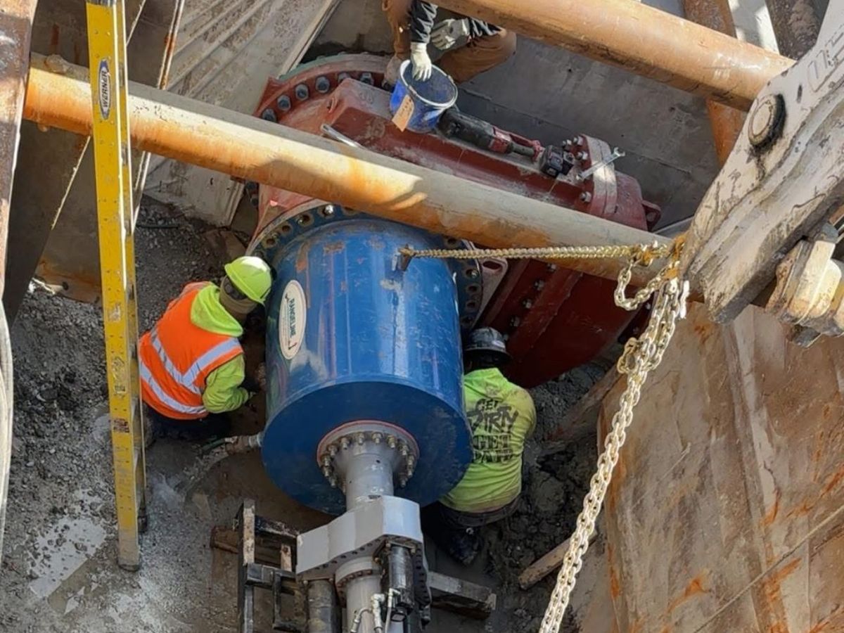 Two JLA & Sons crew members working at a large blue water main fitting and red valve at the bottom of a shored excavation, seen from above.